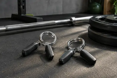 Deux colliers à ressort en acier avec poignées texturées, posés près d’une barre et de disques de musculation.